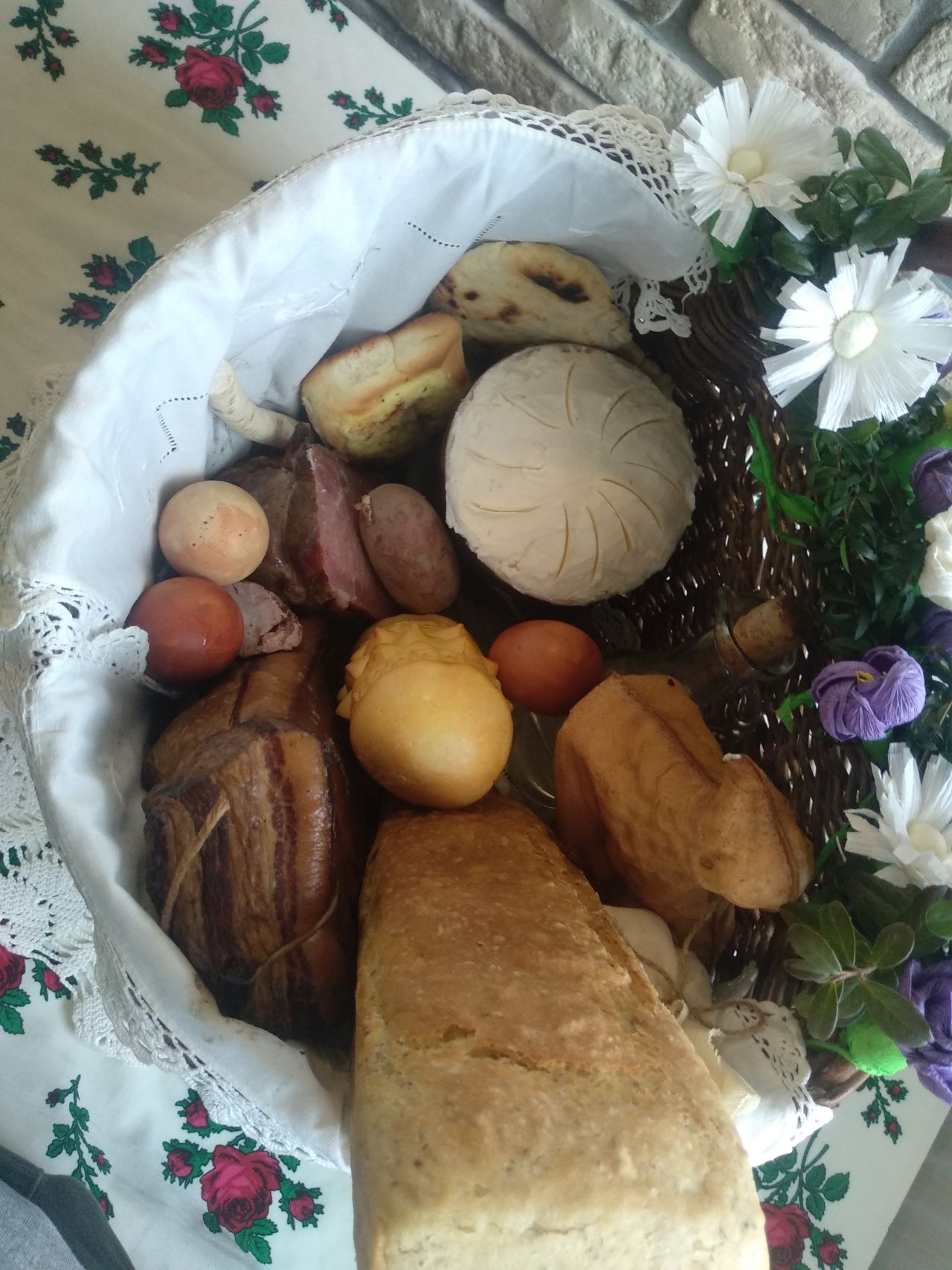 wnętrze wiklinowego koszyka z góry, w środku na białej serwecie chleb z formy, sery, baranek, jajka barwione na ciemnoczerwono, wędliny i inne wiktuały 
