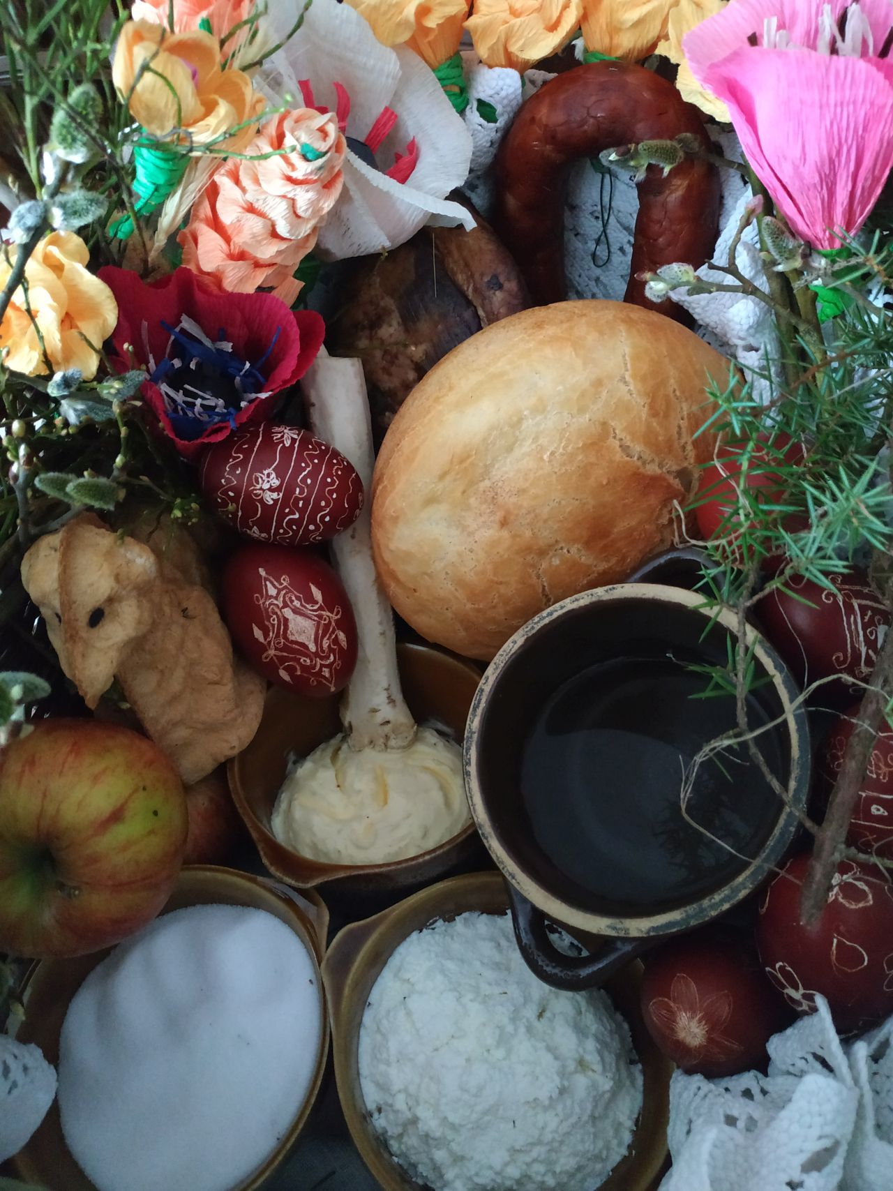 wnętrze koszyka z góry, w środku chleb, baranek, pisanki, kiełbasa, boczek, jabłko, naczynia z masłem, wodą, solą i serem, wokół są kwiaty z bibułył